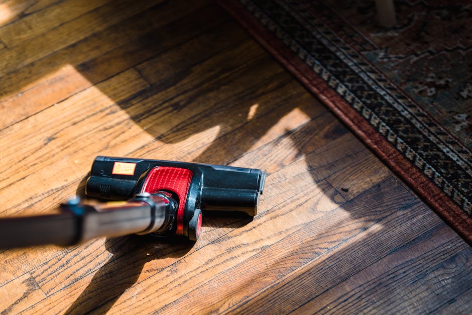 Close-up image of a cordless handheld vacuum cleaner with a black and red body lying on a polished wooden floor near a patterned area rug. The surface of the hardwood floor shows visible wood grain and a clean, shiny appearance, suggesting recent deep cleaning or surface sanitisation. The lighting highlights the natural tones of the wood and casts soft shadows. The corner of the rug, with intricate design and dark red and beige colors, is partially visible, indicating it is placed on the wooden surface in a domestic setting. The image emphasizes the use of modern cleaning tools for thorough surface cleaning, consistent with the residential cleaning services provided by Streatham Carpet Cleaning.