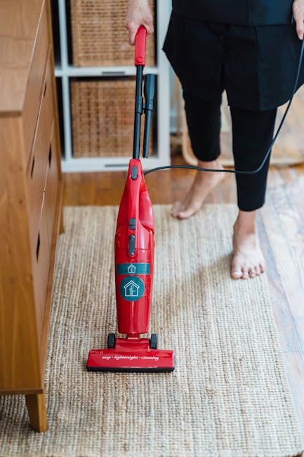 A person, partly visible from the waist down, is vacuuming a beige textured rug in a residential room with wooden flooring and a wooden sideboard. They are wearing black trousers and no shoes, standing on the rug, with a red handheld vacuum cleaner that has a black cord. The vacuum cleaner features a blue power button and a blue house icon on its body. The room is well-lit with natural light, revealing a clean and tidy space that showcases surface cleaning and maintenance typical of domestic cleaning services, as promoted by Streatham Carpet Cleaning for their post-weekend rug care in Streatham Common.