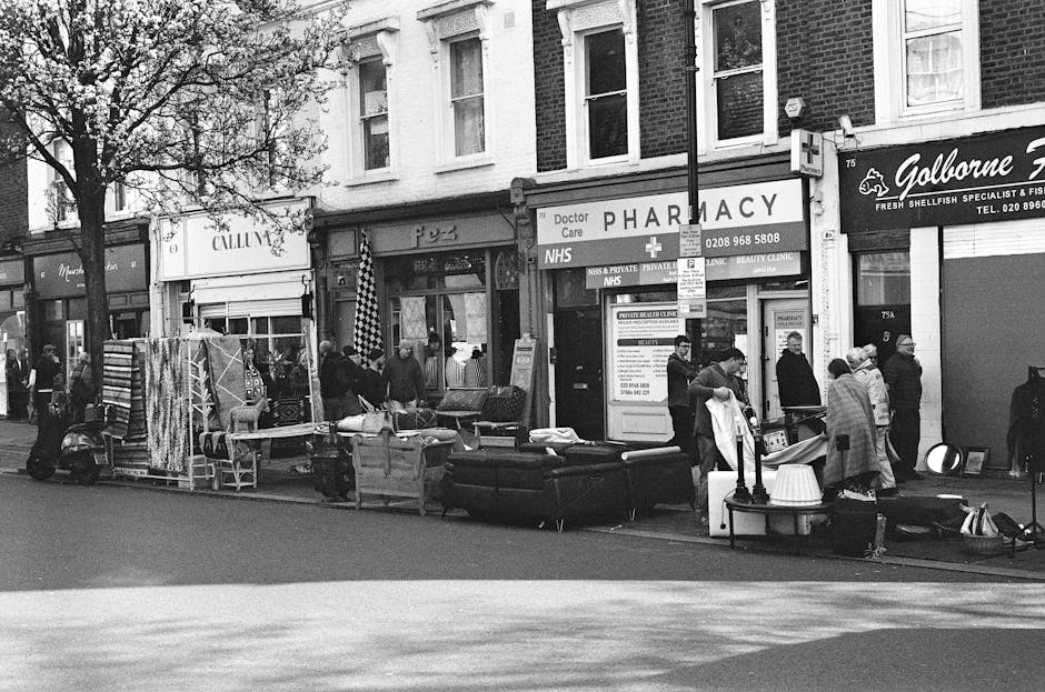 A black-and-white photograph of a busy street scene on Streatham High Road, featuring various storefronts such as a pharmacy and a fish specialist shop. The sidewalk is lined with outdoor displays including chairs, tables, and fabric items, alongside a large sofa with cushions. Several people are browsing and conversing, some wearing coats and hats, indicating cool weather. A streetlamp is centrally positioned, illuminating the scene. In the background, trees with autumn leaves extend over the storefronts, and a scooter is parked at the curb. This image emphasizes the vibrant commercial activity typical of Streatham, with Streatham Carpet Cleaning proudly serving the community, as seen on the nearby signage.