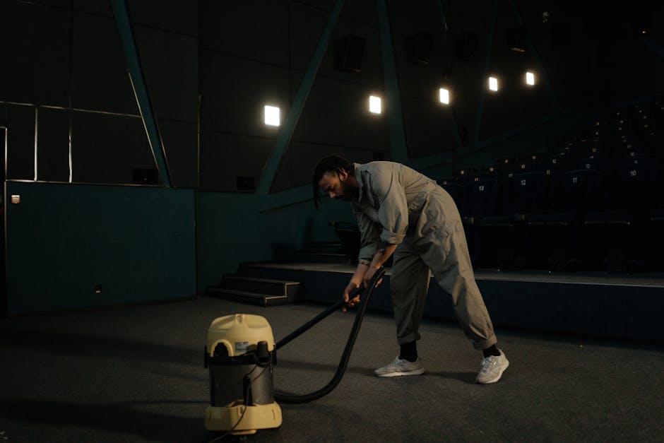 A man in light-colored overalls is performing deep cleaning in an auditorium or theater, using a vacuum cleaner with a long hose attached to a canister-style body. The environment features dark, carpeted flooring and navy blue tiered seating with numbered seats. Overhead, bright rectangular lights illuminate the space, creating a contrast between the illuminated sections and the darker background. The man appears focused on thoroughly cleaning the floor surface, which is free of visible dust or debris, indicating a hygienic, well-maintained space. Streatham Carpet Cleaning offers professional surface cleaning and sanitisation services, ensuring a high standard of hygiene for commercial spaces like theaters and auditoriums.