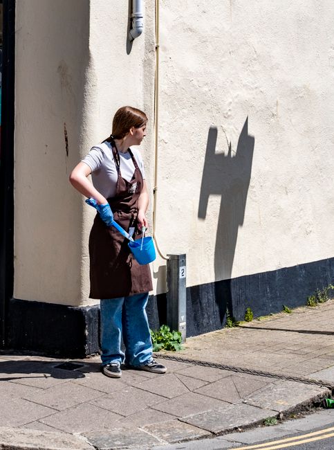 A woman wearing a white t-shirt, a brown apron, and blue jeans is standing outside against a beige wall on a paved sidewalk. She is holding a blue bucket in her left hand and a cleaning spray in her right hand, wearing blue rubber gloves. The scene is illuminated by natural daylight, casting a shadow of a window frame structure on the wall behind her. The area appears clean and well-maintained, with some small green plants growing at the base of the wall. This image represents general domestic or exterior surface cleaning activities, reflecting the services offered by Streatham Carpet Cleaning.